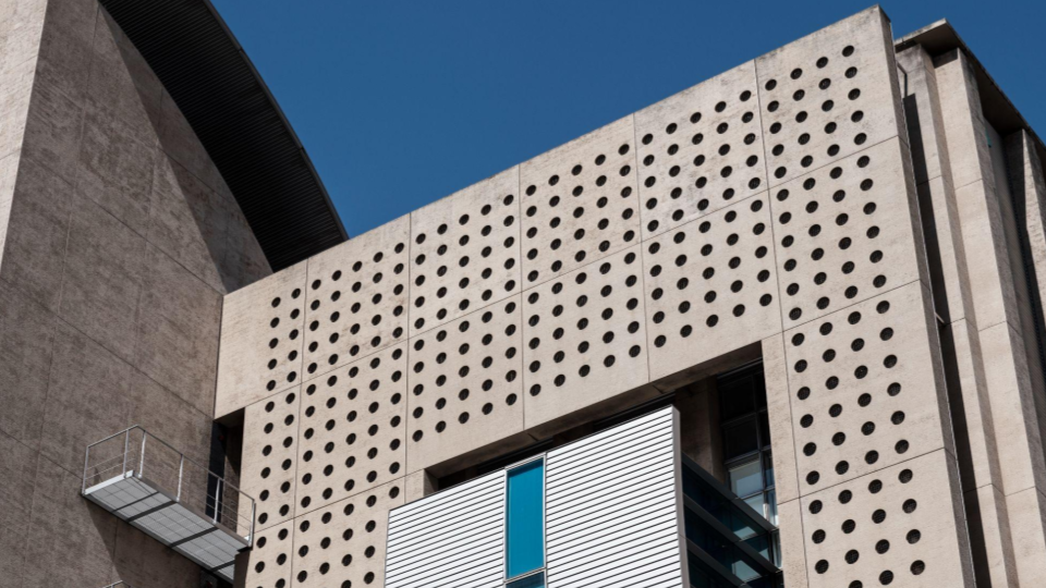 Fachada de um edifício de concreto bruto ou estilo brutalista, repleta de perfurações circulares padronizadas que funcionam como elementos vazados de iluminação.