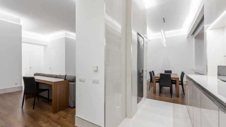 Visão ampla de um ambiente residencial moderno com iluminação embutida no teto, integrando a cozinha, uma área de jantar e uma pequena bancada de trabalho ao fundo.