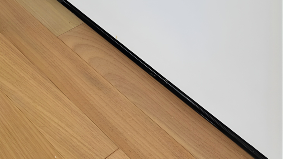 Detalhe em plano fechado da transição entre um piso laminado de madeira clara e uma parede branca, divididos por um perfil ou rodapé fino preto.
