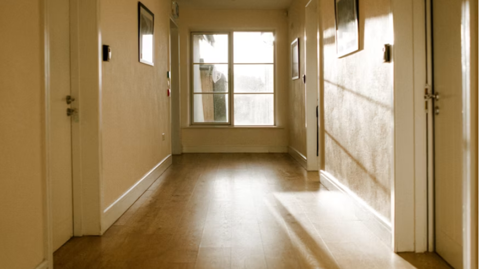Um corredor residencial iluminado pelo sol, com piso de madeira, paredes claras e rodapés brancos clássicos de sobrepor.