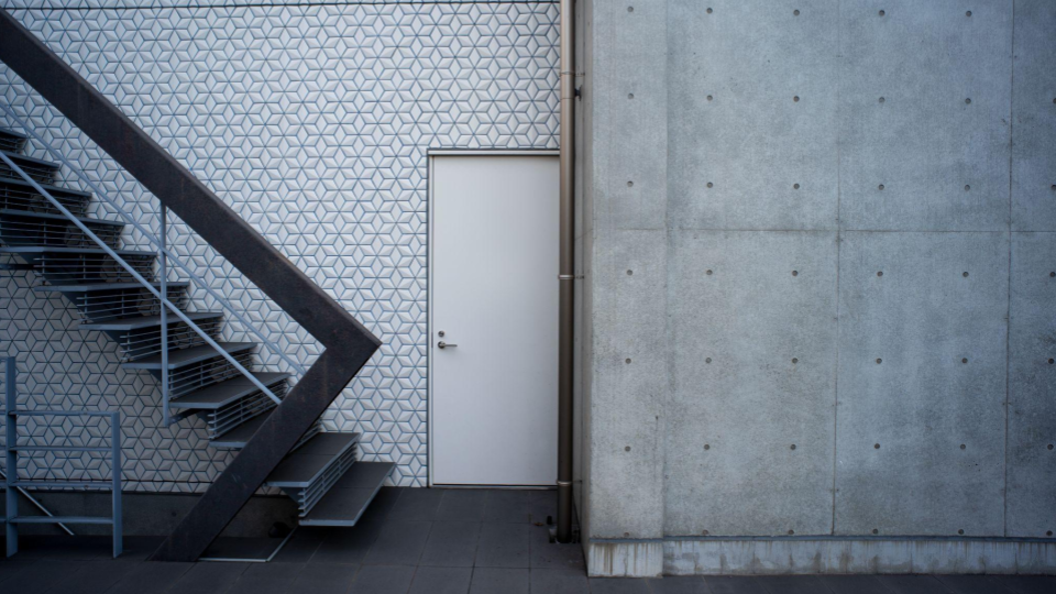 Fachada arquitetônica moderna e minimalista com uma escada externa de metal, uma porta branca centralizada e uma parede de concreto aparente.