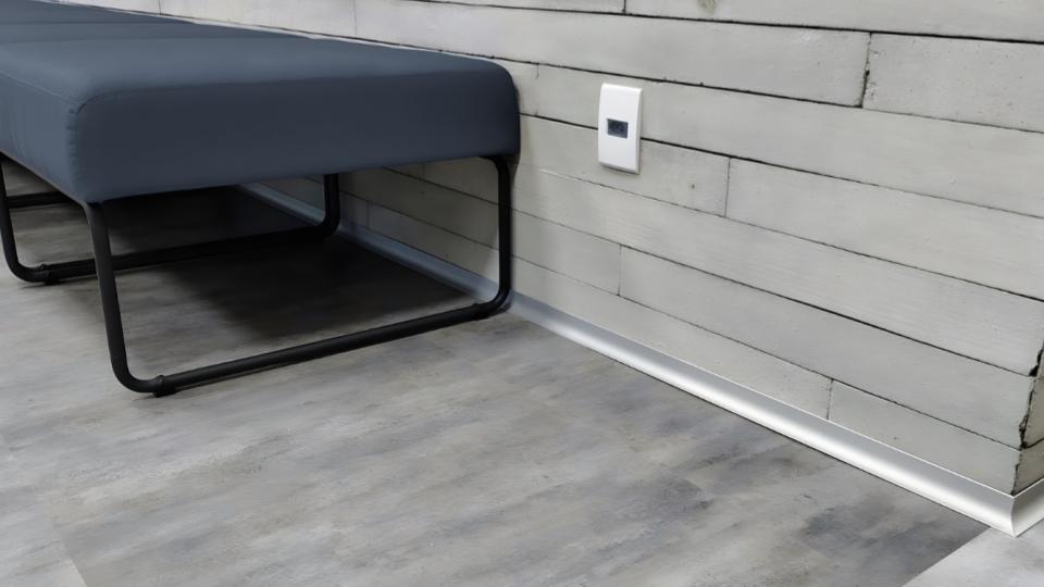 A extremidade inferior de um ambiente com piso em tom cinza-cimento, parede de réguas de madeira clara, uma tomada e o detalhe da instalação de um rodapé metálico/plástico discreto ao lado de um banco de metal e estofado escuro.