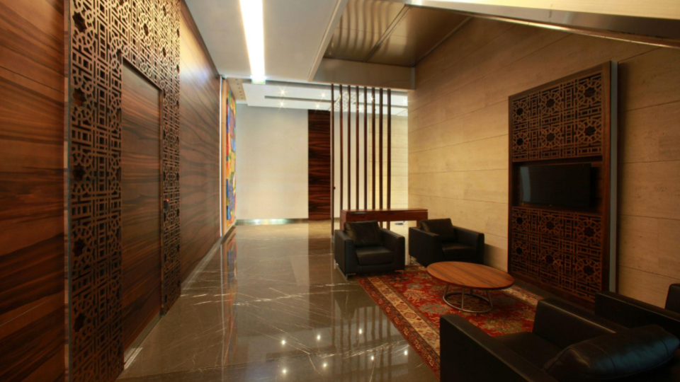 A imagem apresenta a recepção ou hall de entrada de um edifício, decorado em estilo contemporâneo com tons neutros e amadeirados.