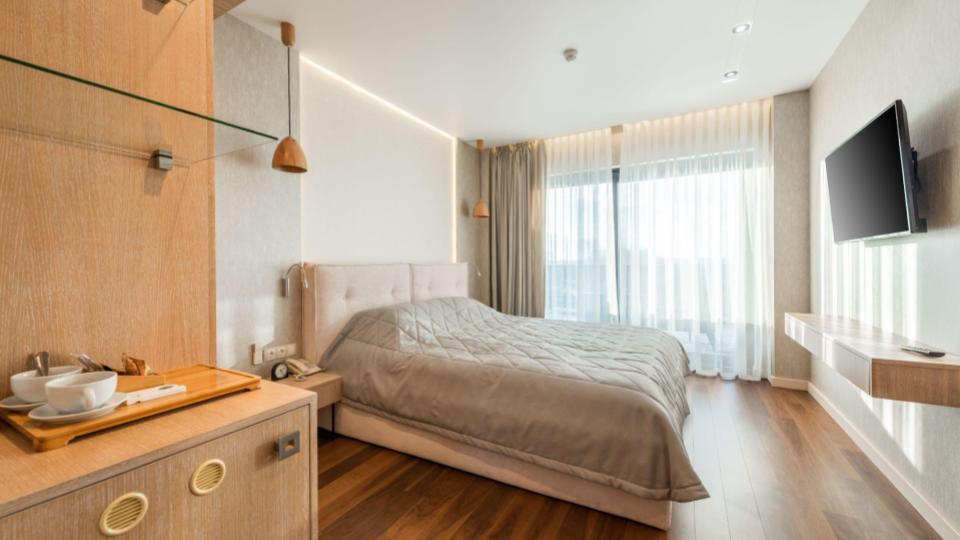 Suíte de hotel bem iluminada, equipada com uma cama de casal com cabeceira estofada e colcha cinza, piso de madeira clara e uma parede de vidro com cortinas translúcidas. Em primeiro plano, há um móvel de madeira com um jogo de xícaras de café.