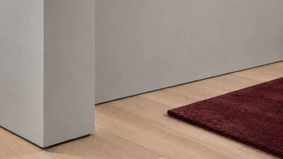Composição de design minimalista focada no encontro de paredes cinza-claras com rodapé embutido discreto, piso de madeira clara e a ponta de um tapete felpudo na cor vinho/bordô.