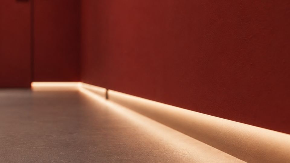 Plano detalhado do encontro de uma parede vermelha com o piso de cimento queimado, destacando um rodapé com fita de LED embutida que projeta uma luz quente e contínua ao longo do chão.