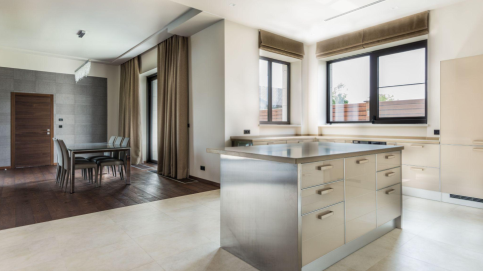 Cozinha com conceito aberto integrada à sala de jantar. Apresenta uma ilha central com acabamento metálico (inox) e armários em tom creme/bege, acompanhada de janelas amplas com persianas romanas e piso de porcelanato claro que transiciona para madeira na área da mesa de jantar.