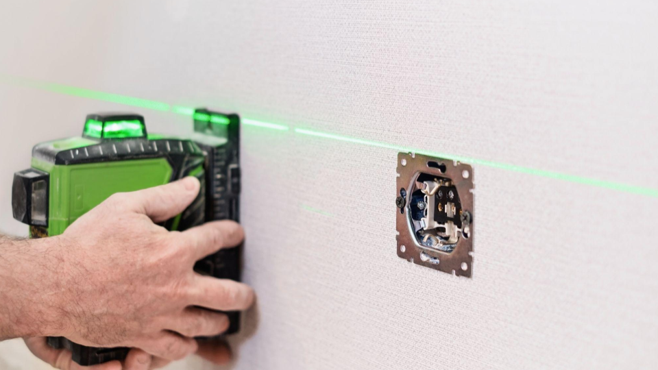 Um profissional segurando um nível a laser verde posicionado na parede para alinhar a altura de uma caixa de tomada/interruptor elétrica de embutir.
