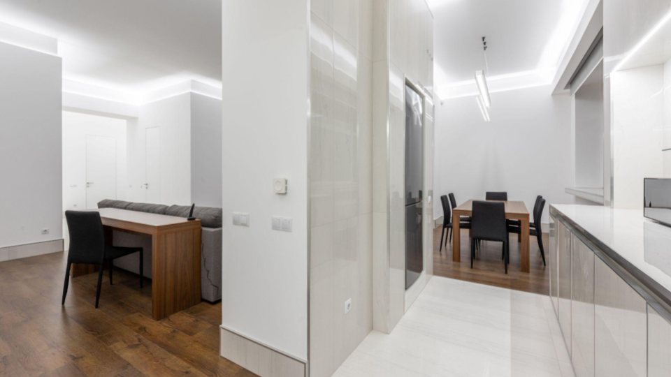Visão ampla de um ambiente integrado (sala, jantar e cozinha) com iluminação no sanca de gesso. O espaço destaca a transição de pisos: madeira escura na área de estar/jantar e porcelanato/mármore claro na área da cozinha.