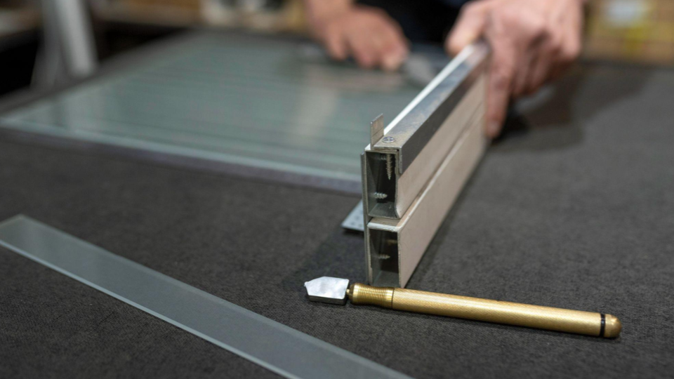 A imagem em plano detalhe mostra o corte de perfil em caixilho/perfil de alumínio instalado sobre um móvel ou painel. No primeiro plano, sobre uma bancada escura, há um cortador de vidro/riscador manual com cabo de latão recartilhado e uma régua ou lâmina de vidro.