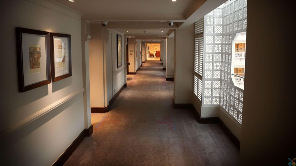 Corredor longo com iluminação amarelada e carpete estampado. As paredes claras exibem quadros decorativos emoldurados e uma das laterais apresenta um painel trabalhado com elementos vazados em estilo oriental/árabe.