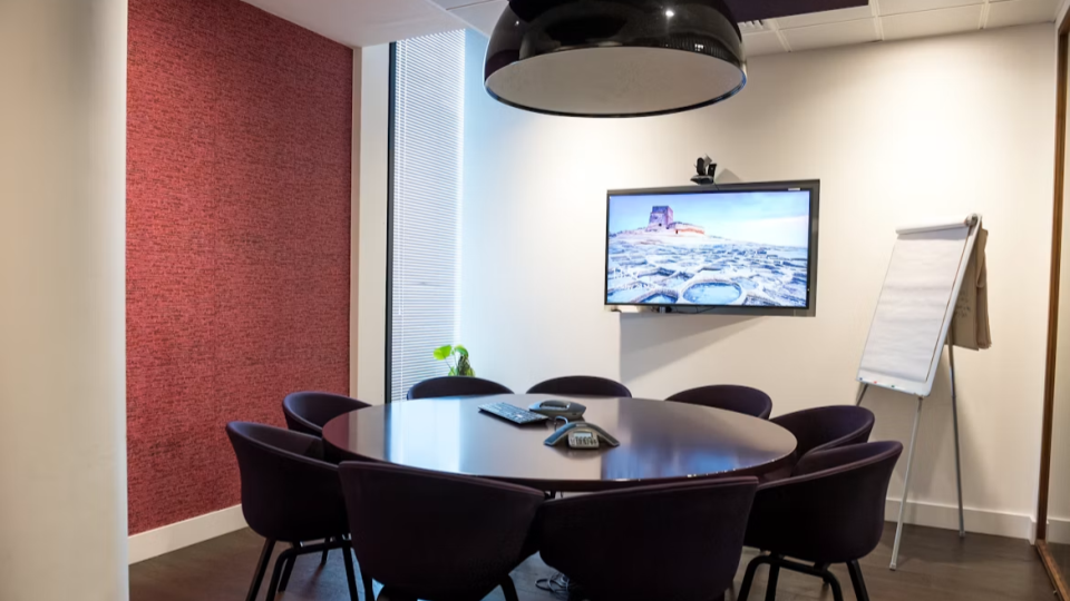 A imagem mostra uma sala de reunião corporativa moderna e elegante, com uma mesa oval de madeira escura cercada por várias cadeiras estofadas em tom vinho ou marrom-escuro.