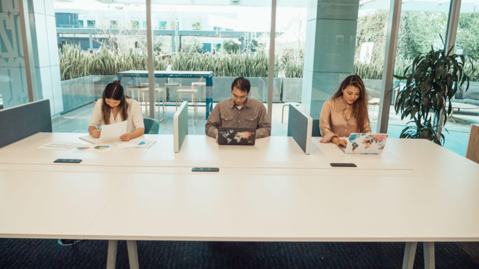 Estações de trabalho em bancada compartilhada (Coworking)
Três pessoas trabalhando lado a lado em uma mesa branca com divisórias acústicas centrais e grande fachada envidraçada ao fundo.