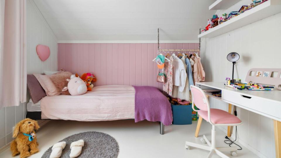 Quarto infantil aconchegante com cama arrumada em tons de rosa e roxo, arara de roupas suspensa, escrivaninha branca e tapete redondo no chão.