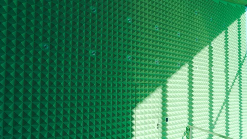 Close em um revestimento acústico verde com textura de pirâmides (cunhas anecóicas), cobrindo uma parede inteira com reflexos de luz e sombra.