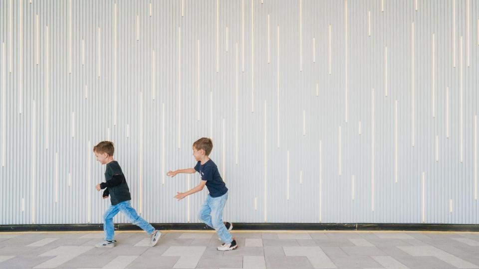 Dois meninos usando calça jeans e camiseta brincam de correr em um corredor com parede ripada iluminada ao fundo.
