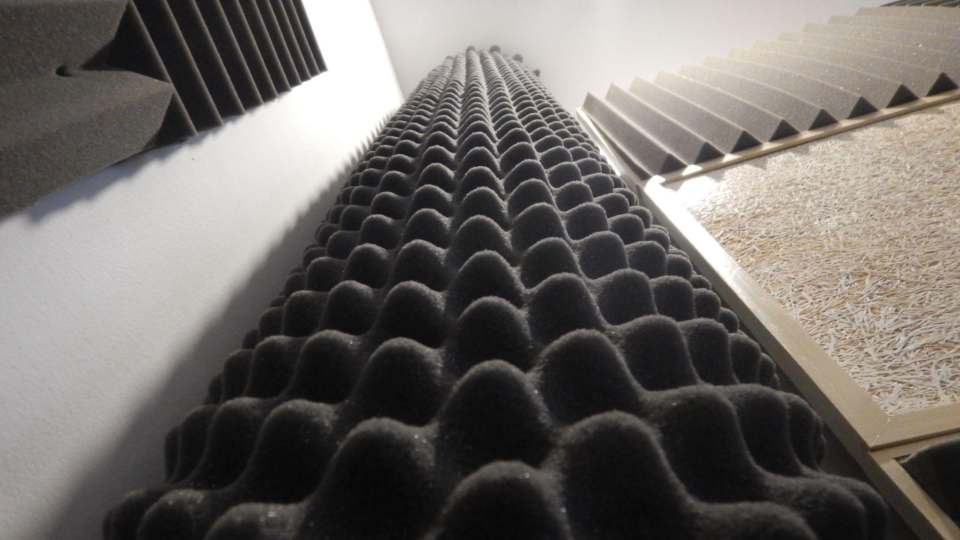 Perspectiva em ângulo baixo destacando diferentes painéis e espumas acústicas. No centro, sobressai uma espuma preta do tipo "caixa de ovo", cercada por placas piramidais e um painel de fibra de madeira.