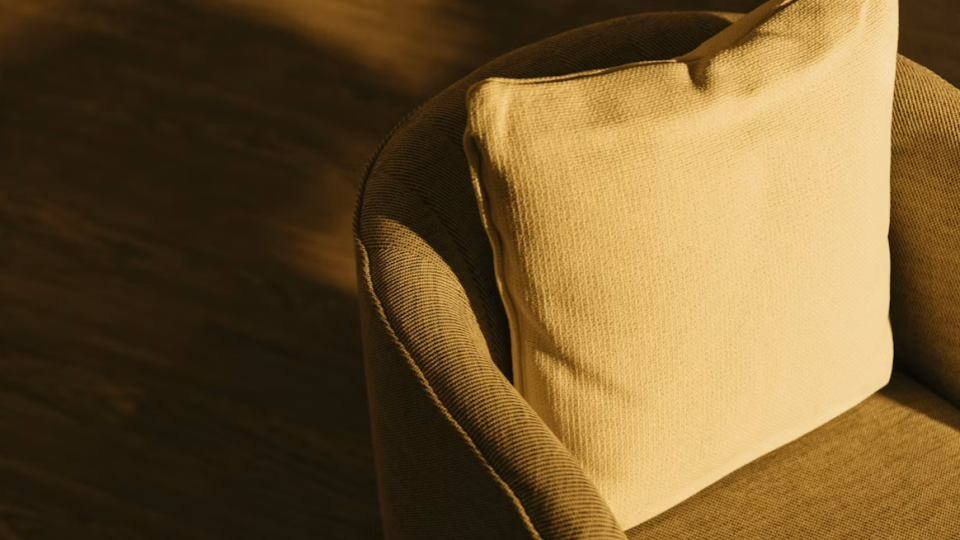 Um close aconchegante do encosto de uma poltrona estofada bege com uma almofada quadrada de tecido texturizado, banhada por uma luz dourada e calorosa.