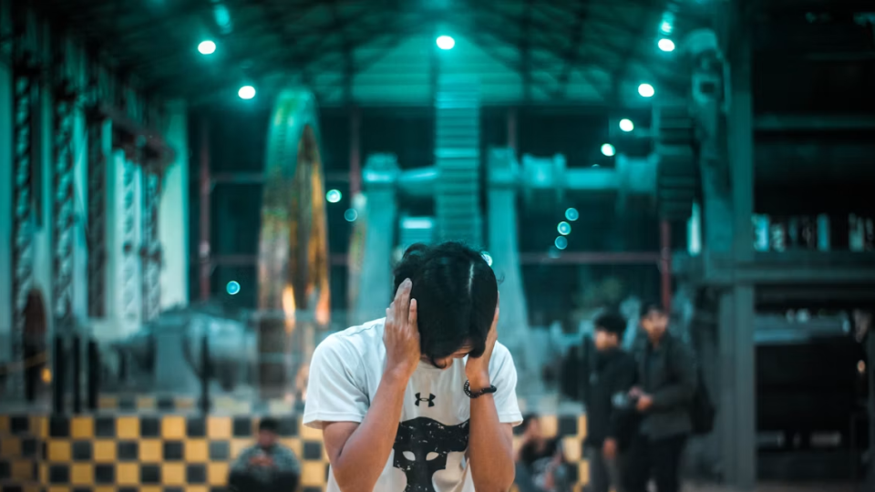 Um rapaz vestindo camiseta branca cobrindo os ouvidos com as mãos em um ambiente industrial/fábrica com grandes engrenagens ao fundo e iluminação esverdeada, sugerindo incômodo com ruído excessivo.