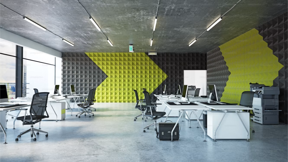 Escritório moderno de estilo industrial com teto de concreto aparente e calhas de iluminação. A parede ao fundo apresenta revestimento acústico piramidal preto e amarelo-esverdeado formando o desenho de uma grande seta. O espaço conta com várias bancadas de trabalho brancas, cadeiras giratórias pretas e uma impressora multifuncional.