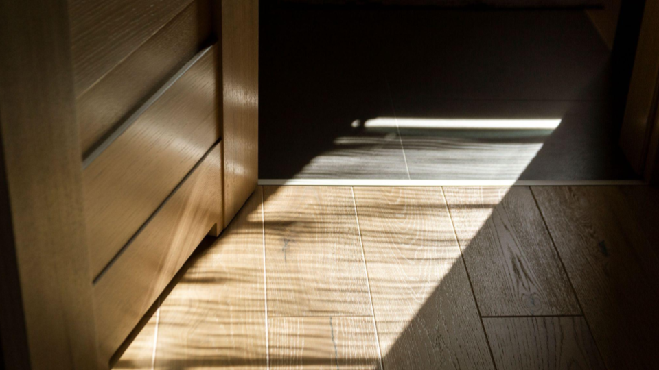 Foto em plano detalhe do piso de um ambiente interno com foco em uma porta de madeira clara parcialmente aberta.